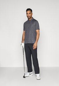 Polo gris avec un motif subtil, associé à un pantalon noir. Le joueur tient un club de golf, portant un gant blanc et des chaussures blanches Nike.