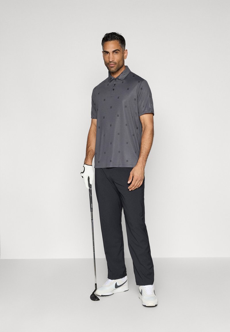 Graues Poloshirt mit subtilen Mustern, kombiniert mit schwarzen Hosen. Der Spieler hält einen Golfschläger, trägt einen weißen Handschuh und weiße Nike-Schuhe.