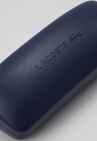 Tamsiai mėlynas kietas dėklas su tekstūruotu paviršiumi, ant viršutinės dalies esantis įspaustas "LACOSTE" logotipas ir krokodilo emblema.