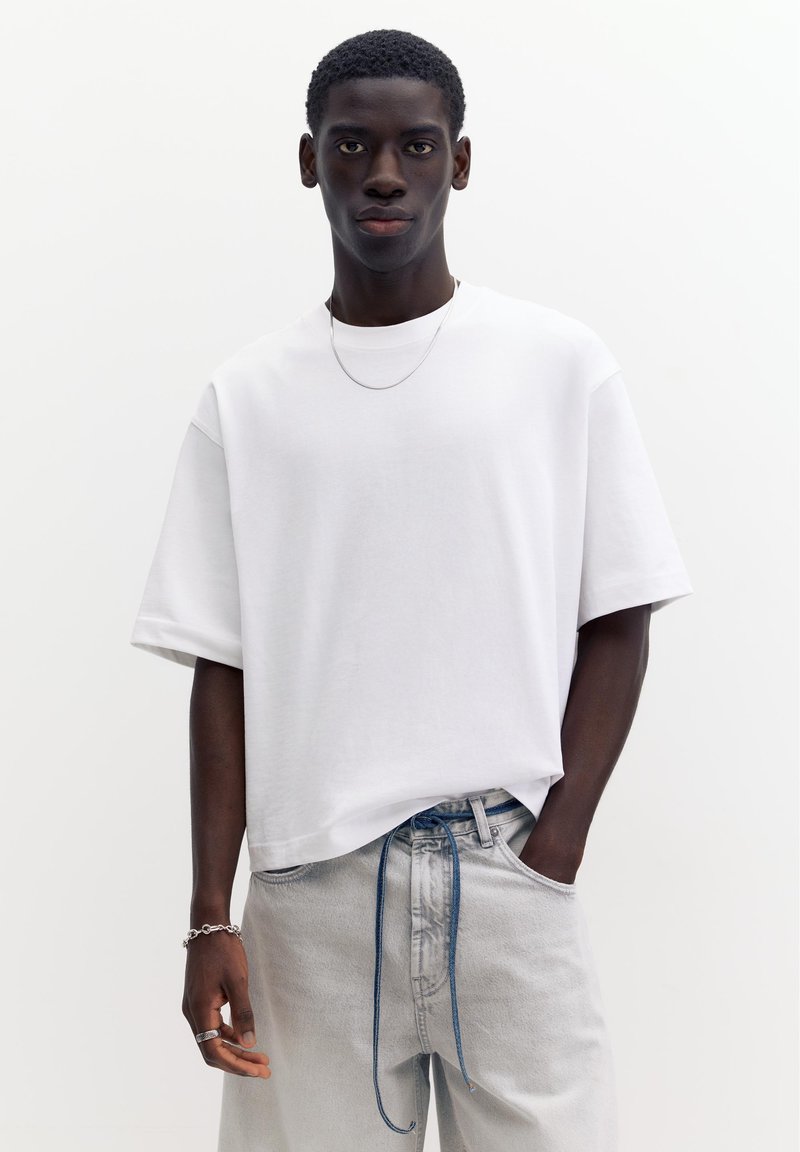 Jeune homme portant un t-shirt blanc ample, un jean gris clair avec un cordon bleu et des bijoux argentés, se tient devant un fond blanc uni.