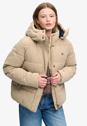 Ung kvinde, der lynlås en beige corduroy dunjakke med hætte, iført blå jeans og en sweater under.