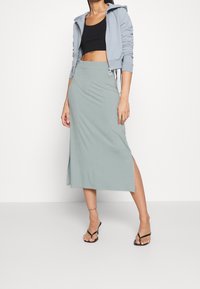 Lyseblå lynlåsjakke, sort crop top og et langt grønt nederdel med en sideslids, parret med sorte flip-flops. Glat, blødt stof.