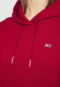Sudadera roja con capucha y cordones con puntas de metal. Cuenta con un pequeño logo bordado en el pecho. Está hecha de una tela suave y texturizada.
