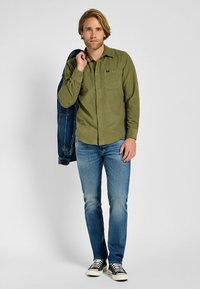 Camisa de botón verde oliva con un bolsillo en el pecho, combinada con jeans azules y zapatillas negras y blancas. Chaqueta de denim caída sobre el hombro.