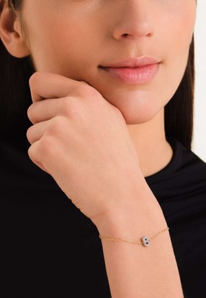 Pulsera de cadena dorada con un charm ovalado central que tiene una piedra preciosa transparente rodeada de piedras más pequeñas, textura suave, diseño elegante.