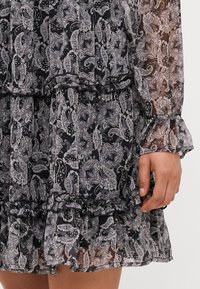 Sorte og hvite paisley-mønstrete kjole laget av gjennomsiktig stoff, med volanger og lange ermer med elastiske mansjetter.