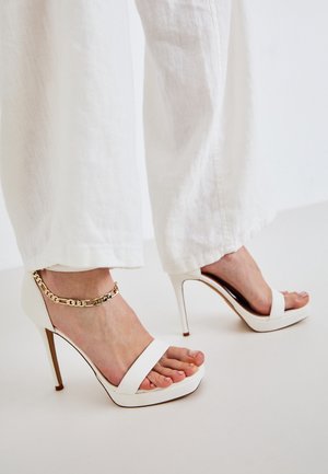 Pieds portant des sandales blanches à talons hauts avec une bride de cheville en chaîne dorée, associées à un pantalon blanc ample.