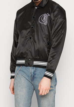 Czarna bomberka z satyny z białymi paskami na mankietach i u dołu, haftowanym logo na piersi, noszona z jasnymi, przetartymi jeansami i łańcuszkowym dodatkiem.