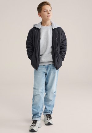 Melna šūta jaka ar pelēku kapuci, gaiši pelēka sporta džemperis, gaiši zilas džinsa bikses un pelēki sporta apavi. Uzsvars uz vairākslāņu faktūrām un ikdienišķu piegriezumu.