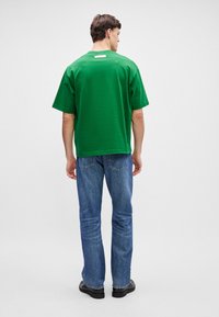 T-shirt de algodão verde com um corte descontraído, mangas curtas e um rótulo nas costas, combinado com jeans de ganga azuis e sapatos pretos.