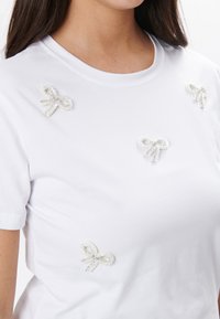 Camiseta de algodón blanca con cuatro aplicaciones decorativas de lazo en tonos pastel con acentos de cuentas, colocadas de manera asimétrica en la parte frontal.