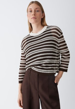 Vrouw met steil blond haar, gekleed in een bruin-wit gestreepte gebreide trui en donkerbruine broek, staand met één hand in haar zak.