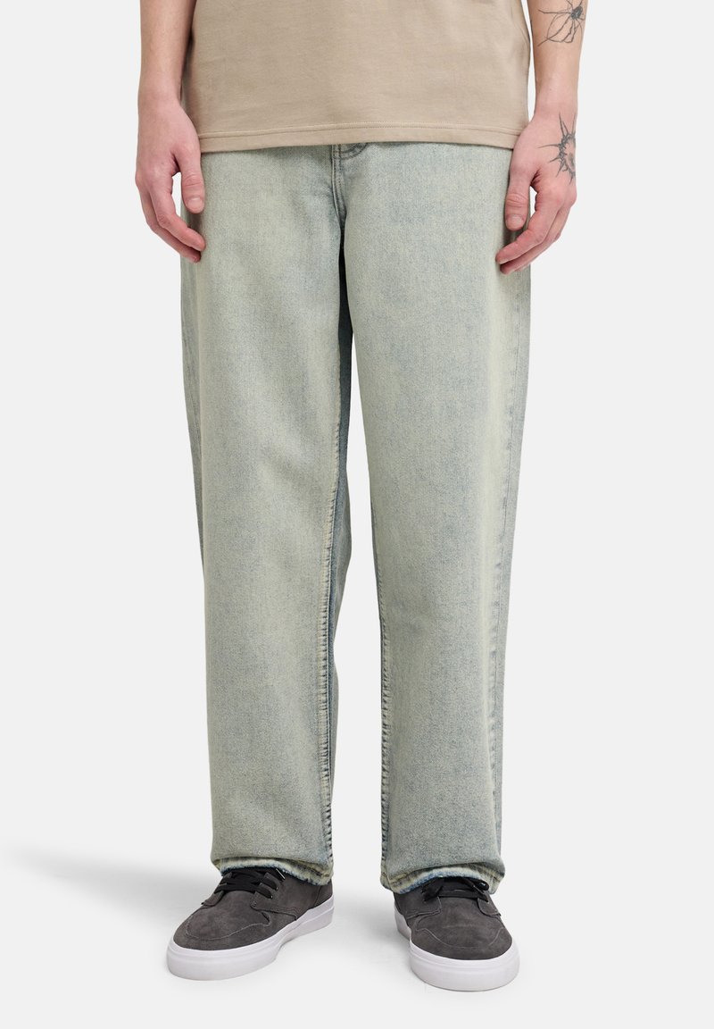 Μπλε ξεβαμμένο τζιν με χαλαρή εφαρμογή, ίσια μπατζάκια και διακριτικές λεπτομέρειες υφής, συνδυασμένο με γκρι αθλητικά παπούτσια.