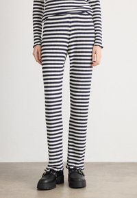 Pantalone od rebraste tkanine u tamnoplavim i bijelim prugama. Ima uski kroj i opušteni fit sa volanima na rukavima. Upareni s crnim cipelama.