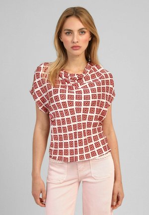Jeune femme aux cheveux blonds porte un chemisier rouge et blanc à manches courtes à motif et un pantalon rose clair sur un fond gris uni.