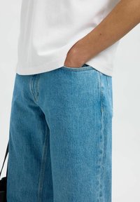 Blue denim jeans with a loose fit, featuring a classic five-pocket design and light stitching details, paired with a white cotton t-shirt.