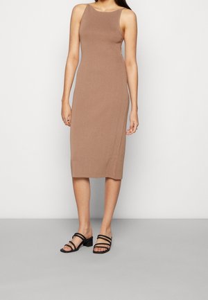 Žena v bezrukávových taupe šatech po kolena s vysokým výstřihem a černými sandály na blokovém podpatku, stojící na šedé podlaze.