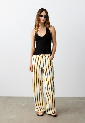 Žena u crnom topu bez rukava, bijelim širokim hlačama s smeđim prugama, sunčanim naočalama i sandalama, stoji s jednom rukom u džepu na sivoj podlozi.