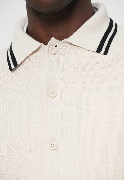 Krēmkrāsas polo krekls ar teksturētu adījumu. Melnbalta svītrainā apkakle, trīs bēši pogas un vertikāla ribota apdare uz ķermeņa.