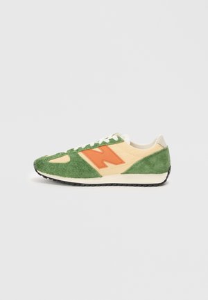 Niskoprofilowy sneaker z zielonego zamszu, beżowymi panelami materiałowymi, pomarańczowym logo "N", białymi sznurowadłami i czarną gumową podeszwą na szarym tle.