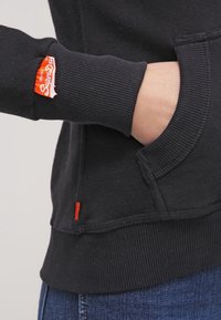 Sudadera negra con puños acanalados y un bolsillo frontal; presenta una pequeña etiqueta naranja en la manga y otra en el dobladillo.