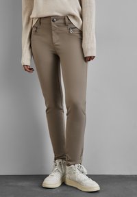 Beige enganliegende Hosen mit Reißverschlusstaschen, kombiniert mit weißen High-Top-Sneakern. Aus weichem, dehnbarem Material und maßgeschneidertem Design.