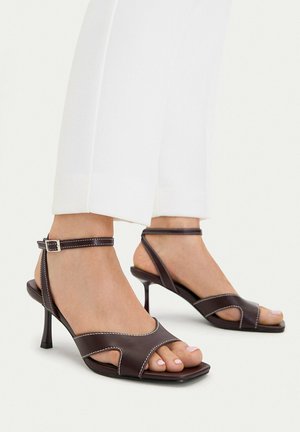 Piedi che indossano sandali con tacco alto in pelle marrone scuro con cinturini alla caviglia e cuciture bianche, abbinati a pantaloni bianchi corti.