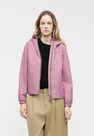 Kvinde med langt blondt hår iført en lyserød hættejakke over en sort top og beige bukser med vide ben foran en ensfarvet hvid baggrund.