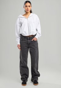 Harmaa lyhyt huppari, jossa on etuvetoketju ja narut, yhdistettynä löysiin tummanharmaisiin farkkuihin. Mustat tennarit täydentävät asua.