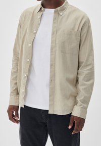 Light beige shirt with a button-down collar, long sleeves, and a chest pocket. Worn over a white t-shirt, paired with dark jeans.