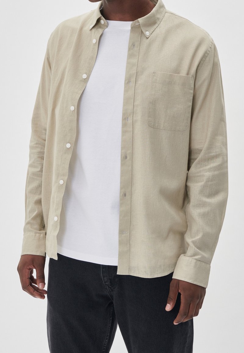 Light beige shirt with a button-down collar, long sleeves, and a chest pocket. Worn over a white t-shirt, paired with dark jeans.