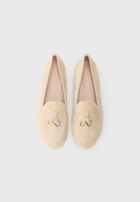 Beige suede loafers with a flat sole, featuring a decorative knot and two-tone metal hardware accents on the front. Smooth inner lining.
