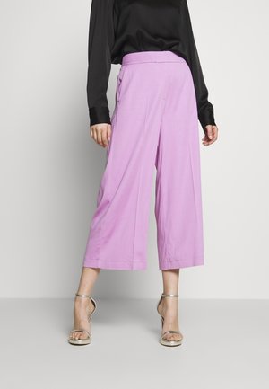 Persoon die lichtpaarse wijd uitlopende cropped broek draagt, een zwart shirt met lange mouwen en zilveren hoge hakken, staand tegen een effen achtergrond.