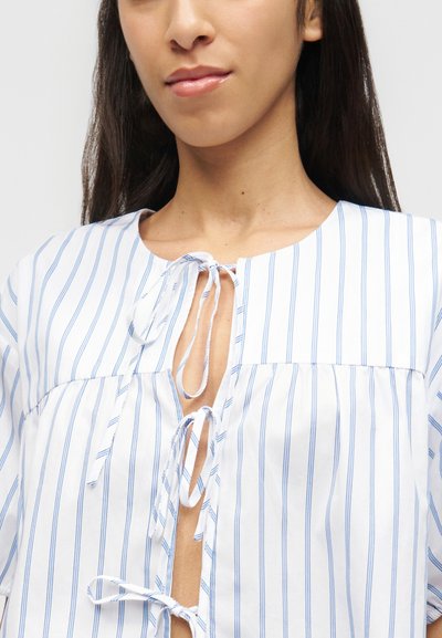 Γυναίκα που φοράει λευκό πουκάμισο με λεπτές μπλε κάθετες ρίγες, με τρία κουμπώματα στο μπροστινό μέρος και στρογγυλή λαιμόκοψη.