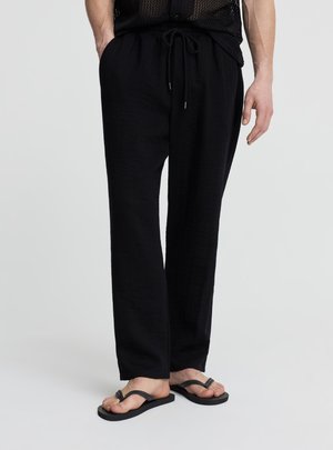 Persona che indossa pantaloni neri con coulisse e infradito nere, in piedi con una mano in tasca davanti a uno sfondo semplice.