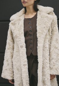 Kvindelig iført en beige faux fur frakke over en mørkebrun knap-top og matchende bukser foran en mørk baggrund.