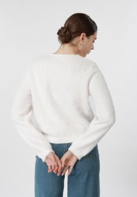 Γυναίκα με τα μαλλιά της πιασμένα σε κότσο, φοράει λευκό χνουδωτό πουλόβερ και μπλε τζιν, στέκεται με τα χέρια δεμένα πίσω, στραμμένη μακριά από την κάμερα.