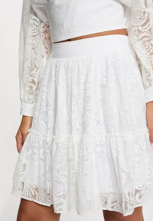 Jupe en dentelle blanche avec motifs floraux et papillons portée sous un crop top assorti à manches longues laissant apparaître un espace au niveau du ventre.