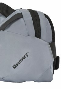 Bolso de hombro de tela gris con acentos negros, que cuenta con una superficie texturizada, compartimentos con cremallera y el logo "Discovery". Diseño compacto y versátil.