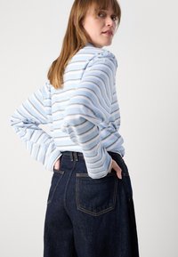 Top a righe blu e bianche con maniche lunghe e ampie, abbinato a jeans in denim blu scuro con tasche posteriori e cuciture in evidenza.