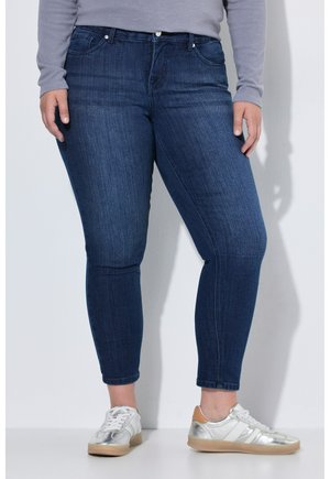 Person iført mørkeblå skinny jeans, grå langærmet bluse og sølvfarvede sneakers, der står op ad en hvid væg.
