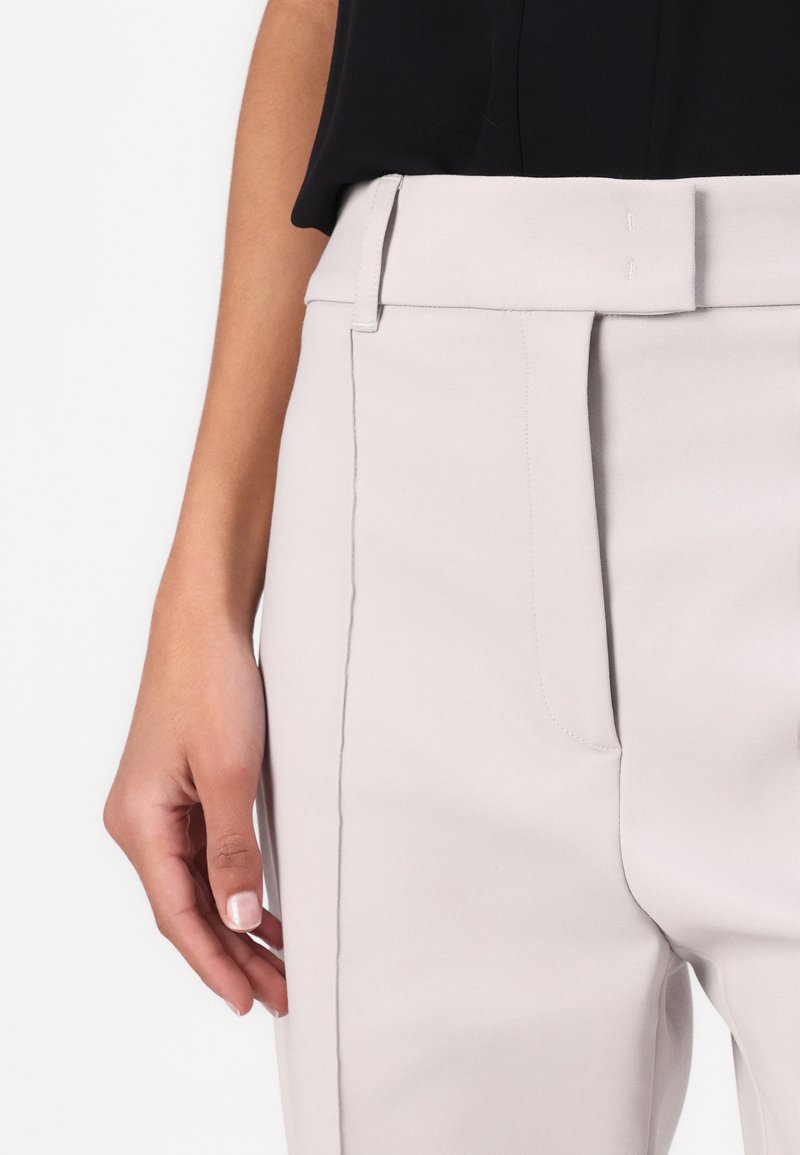 Pantalons ajustés gris clair avec une texture lisse, taille repliée et poches latérales. Main reposant sur la couture latérale.