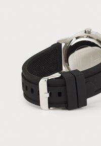 Correa de reloj de goma negra con una superficie texturizada, hebilla de metal plateado y fondo de acero inoxidable, con un diseño de caja hexagonal.