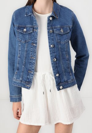 Vrouw draagt blauwe spijkerjack over een witte jurk met koord in de taille, staand tegen een egale lichte achtergrond.