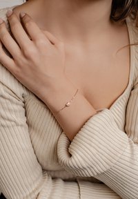 Pulsera de oro rosa con un charm en forma de corazón, diseño de cadena delicada, llevada en un brazo de piel clara. Suéter de crema con textura en el fondo.