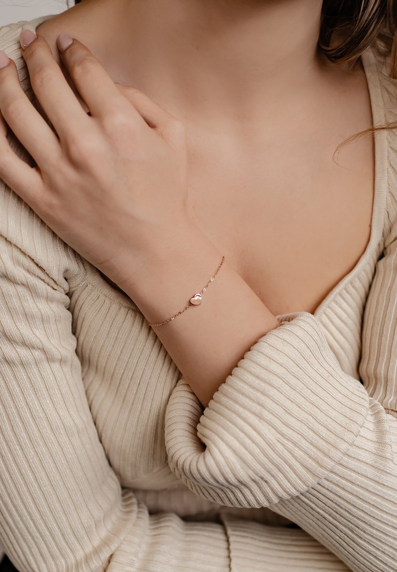 Pulsera de oro rosa con un charm en forma de corazón, diseño de cadena delicada, llevada en un brazo de piel clara. Suéter de crema con textura en el fondo.