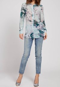 Camicia a maniche lunghe con motivo floreale in tessuto traslucido, caratterizzata da tonalità di blu e verde, abbinata a jeans azzurri chiari e tacchi a righe bianche e nere.