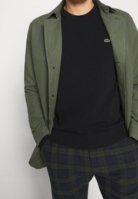 Personne portant une veste vert olive sur un pull noir avec un petit logo crocodile, associée à un pantalon à carreaux vert foncé et bleu marine.