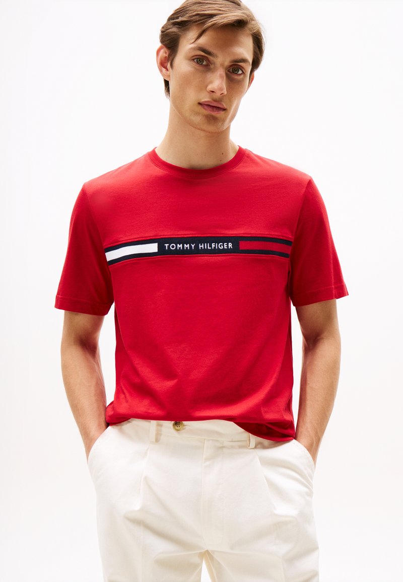 Jeune homme portant un t-shirt rouge Tommy Hilfiger avec des rayures blanches et bleu marine, et un pantalon blanc, debout les mains dans les poches.