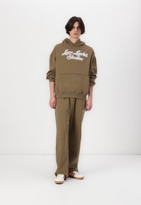Olivgrüner Hoodie mit "Low Lights Studios" in Weiß gestickt, kombiniert mit passenden lockeren Hosen und cremefarbenen Sneakern mit roten Akzenten.